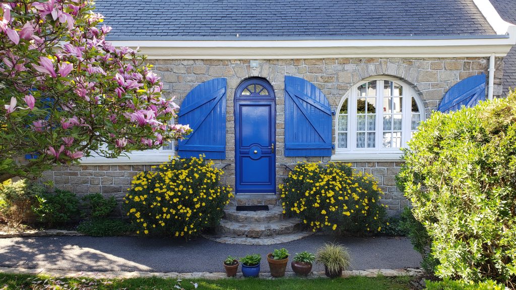 Front facade of a home in Benodet, France with a flowering tree and bushes and ocean blue front door and shutters