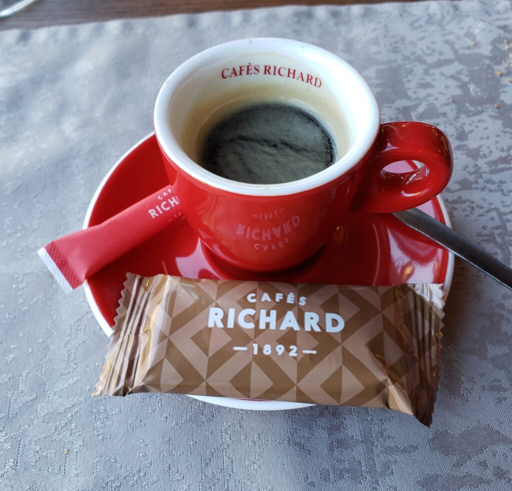 A red "Cafés Richard" espresso cup on a red saucer with coffee in it and a sugar pouch and cookie.