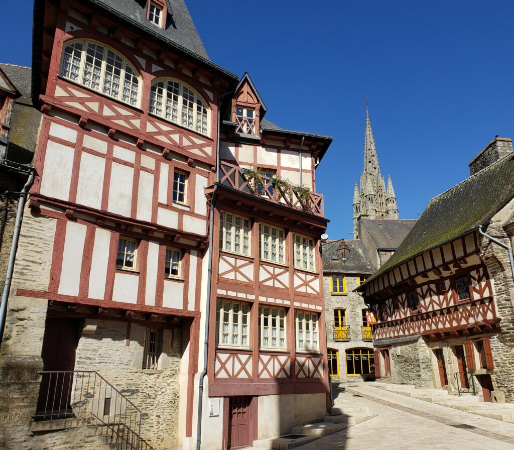 Two half-timbered houses sit on a medieval village street with a gothic church spire behind them.