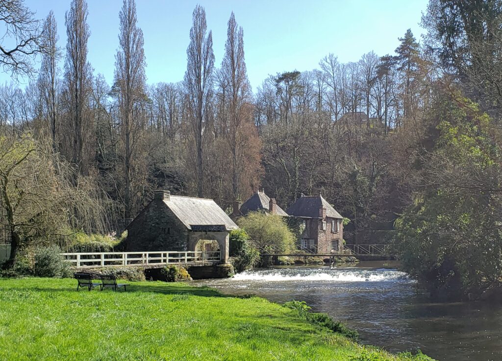 A water mill sits across a river from a grassy park with a low bridge connecting the two. 
